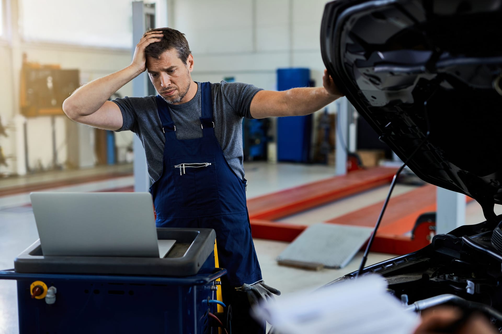 Distraught mechanic having problems while repairing car engine and using laptop in auto repair shop.